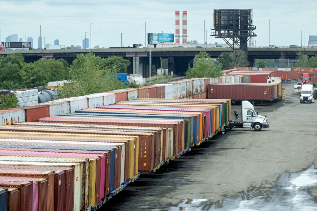 A long row of dozens of differently colored shipping containers are lined up in a large concrete lot under an overcast grey sky. A few semi-truck tractor units are present, with one in the process of being coupled to a shipping container on a trailer. A freeway overpass crowded with cars is in the background. Behind the freeway are two large white-and-red painted smokestacks and the skyline of skyscrapers.
